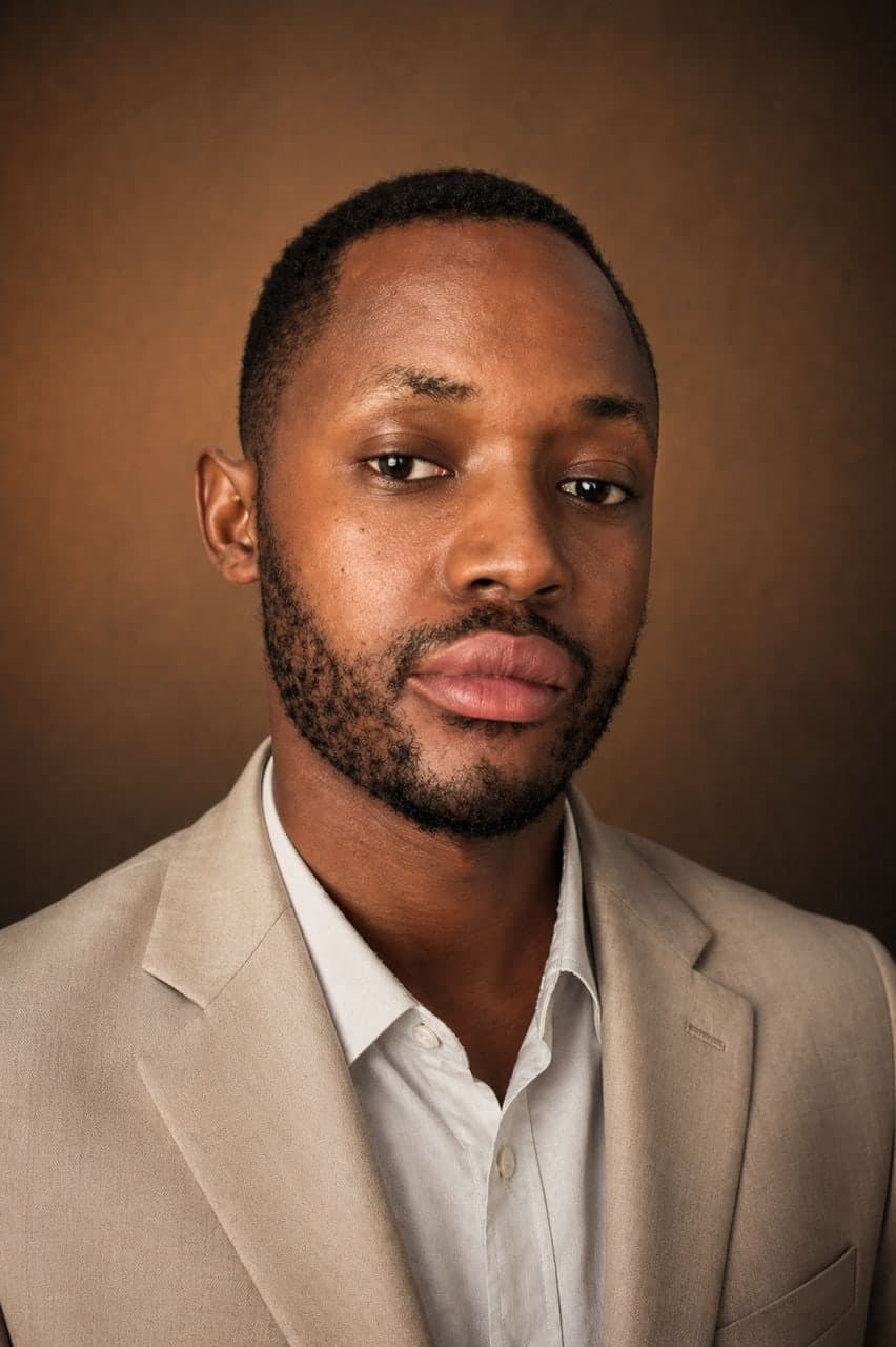 Portrait of Solomon John in a cream suit and white shirt against a warm brown backdrop.
